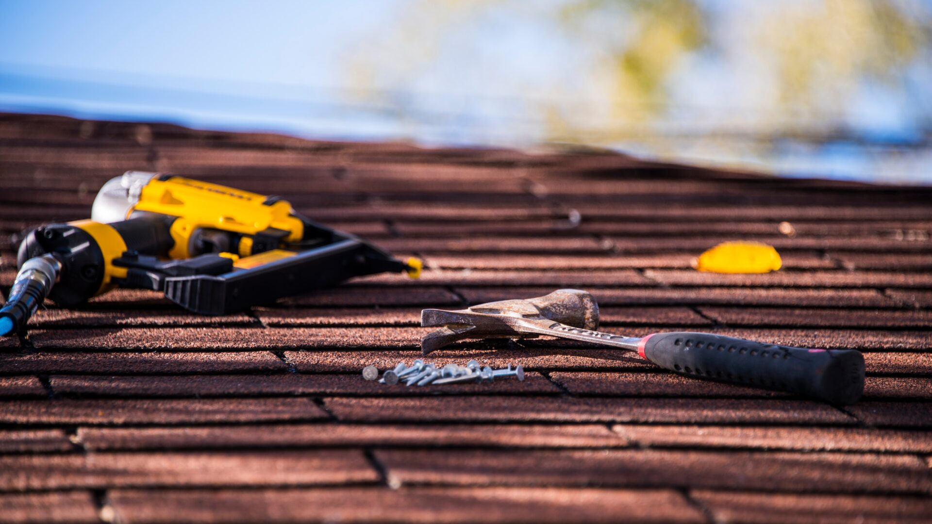 A shingle roof with a hammer, roofing nails, and nail gun sitting on it
